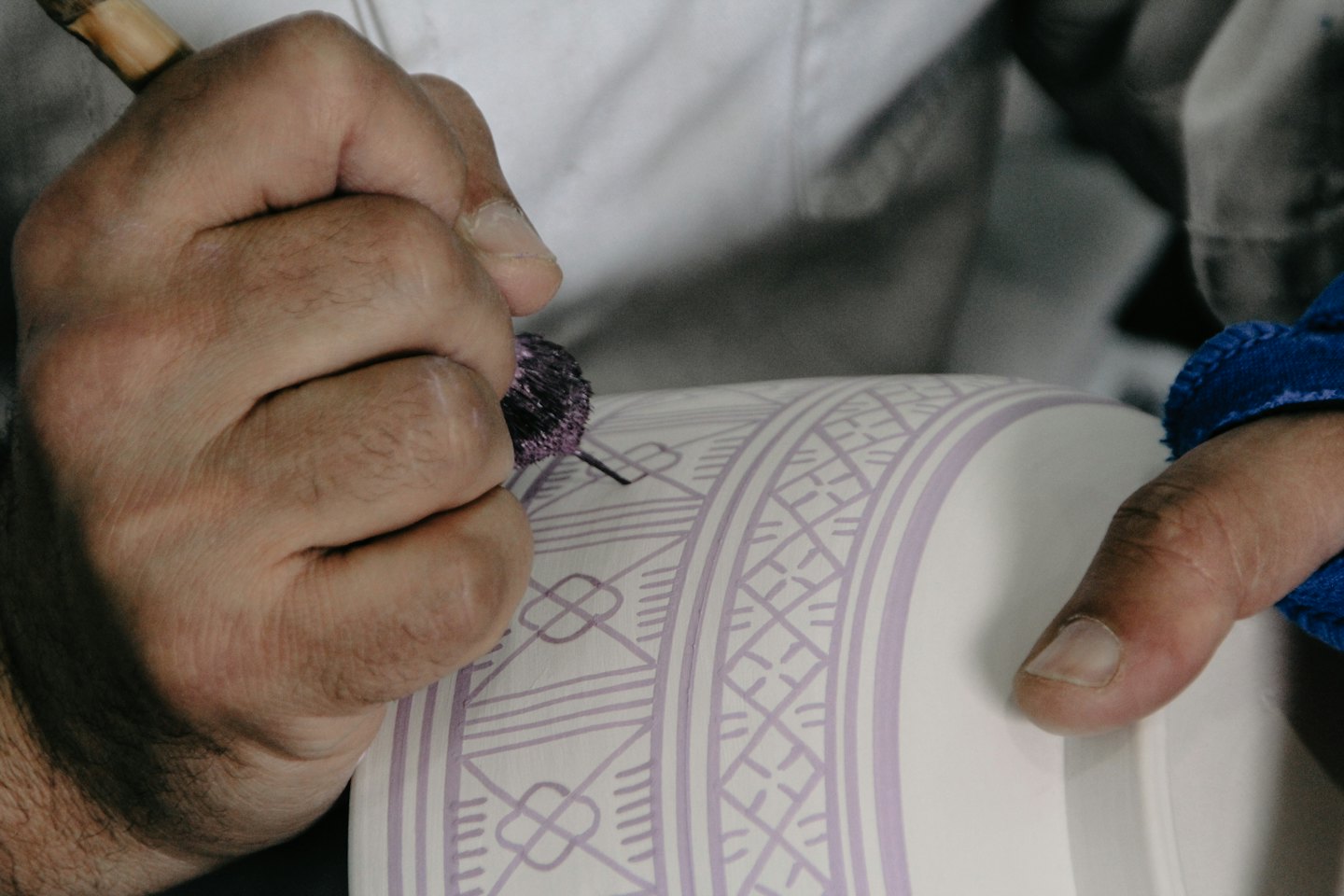 Hände verzieren ein weißes Keramikobjekt mit einem lila geometrischen Muster.